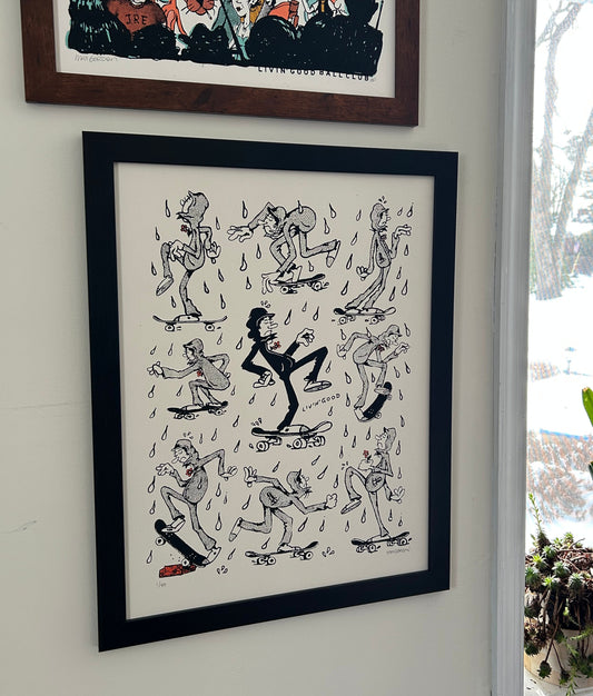 A framed black and white comic-style art print depicting cartoon characters on skateboards in the rain, hanging on a white wall next to a window with potted plants.