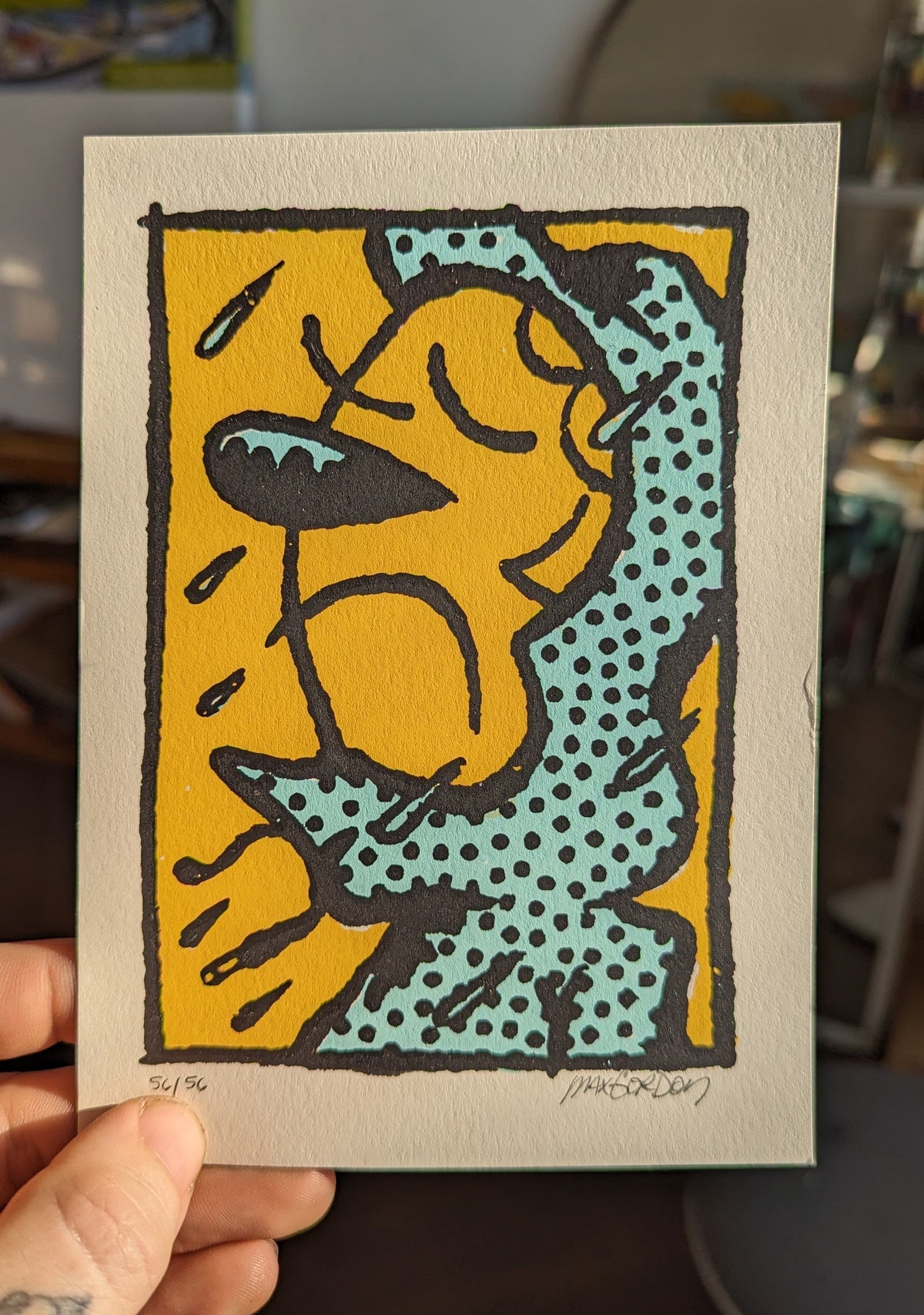 A hand holds a square art print featuring a stylized cartoon cat in a yellow and teal pop-art design with black outlines, photographed in an indoor setting.