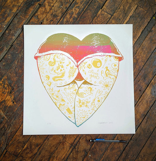 A square art print featuring a stylized heart-shaped face with patterned cheeks and a pink-red top, displayed on a dark wood floor with a pencil below it.