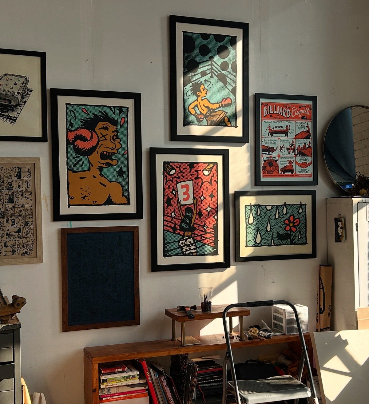 A gallery-style wall display of multiple framed art prints in a real room, with a wooden shelf, ladder, mirror, and sunlight casting shadows across the wall.