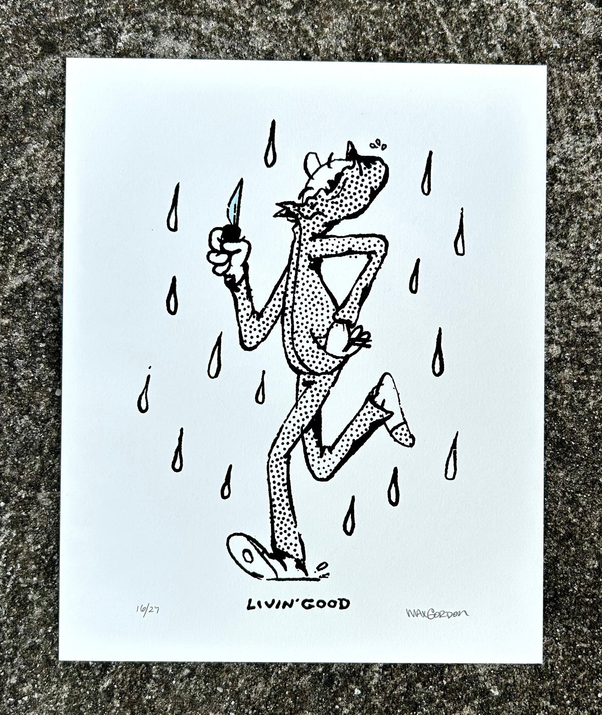 A white art print featuring a black hand-drawn illustration of a cat running in the rain while holding a knife, titled 'LIVIN' GOOD' by Wolfgang Paape, displayed on a textured gray stone surface.