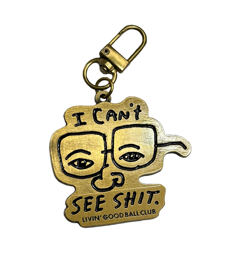 A gold-colored metal keychain featuring a stylized cartoon face with the text 'I CAN'T SEE SHIT. LIVIN' GOOD BALL CLUB' on a plain white background.