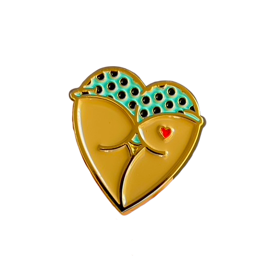 A heart-shaped butt enamel pin in gold-tone metal with a turquoise and black polka-dot patterned booty shorts and a small red heart accent tattoo.