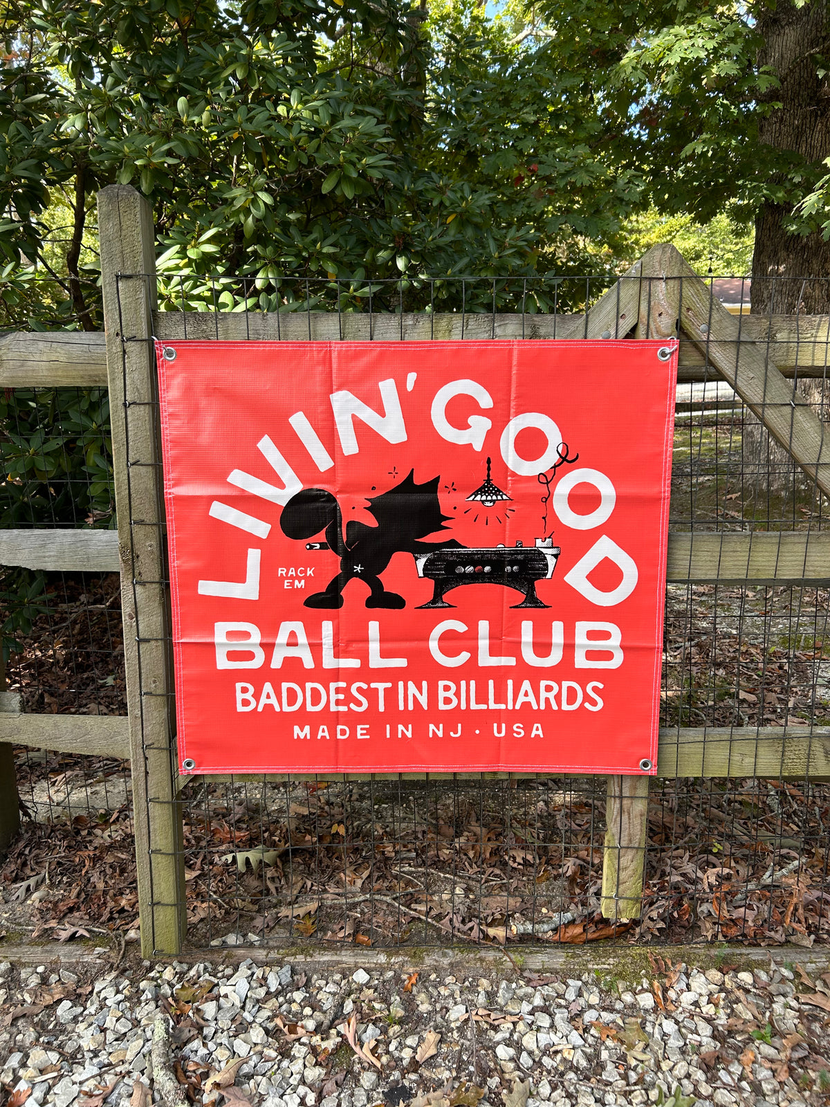 A red rectangular banner hanging on a wooden fence outdoors, with white text reading “LIVIN’GOOD BALL CLUB” and “BADDEST IN BILLIARDS,” plus a black silhouette of a person playing billiards. Trees, fence, and gravel are visible around it.