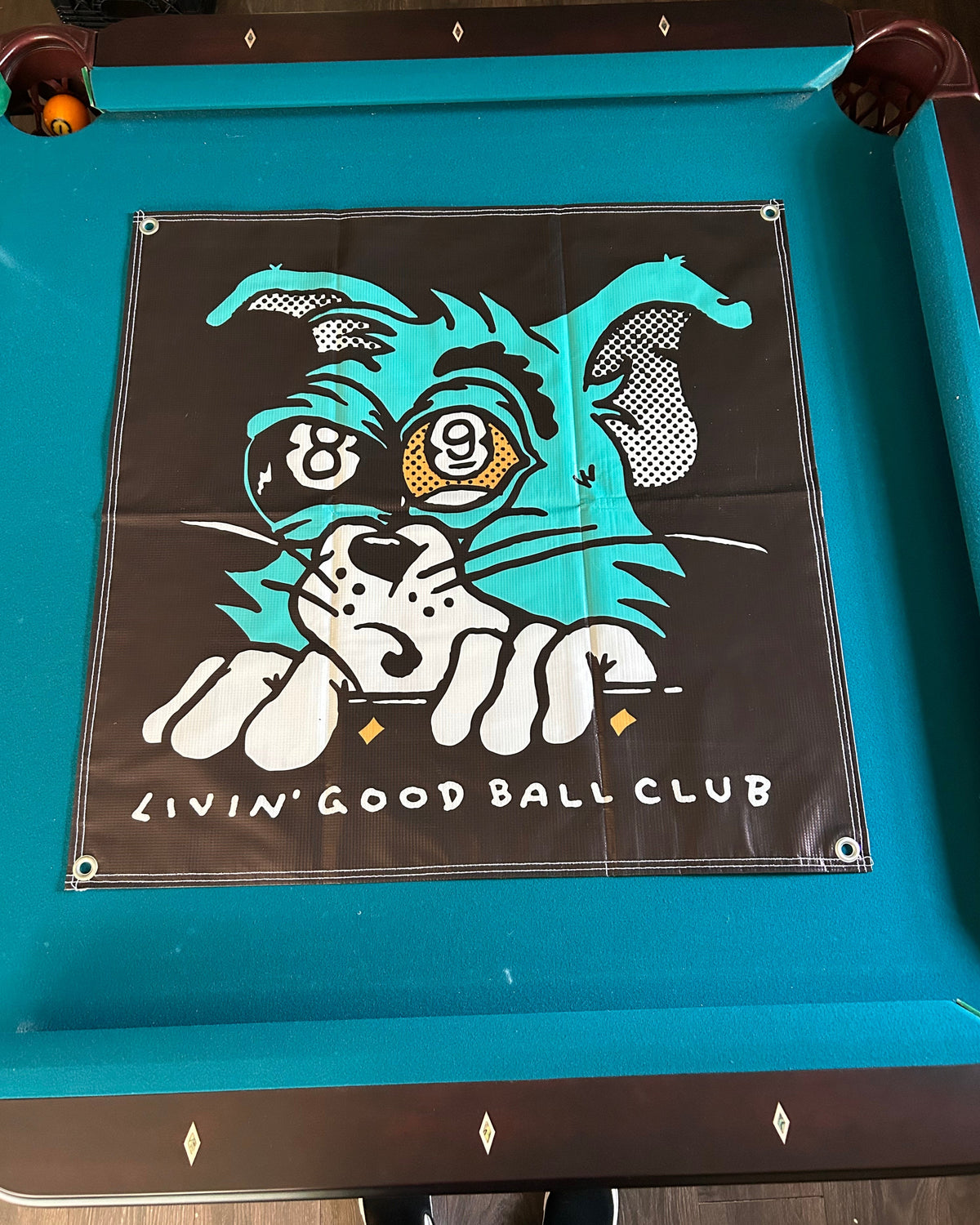 A square black pool table flag featuring a stylized teal, white, and yellow wildcat graphic with the text 'LIVIN' GOOD BALL CLUB' and a nine-ball illustration, displayed on a teal pool table.