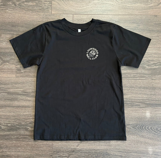 A black short-sleeve t-shirt with a small white circular graphic on the left chest, laid flat on a gray wood-look floor. The graphic contains a skull and the words 'KILLER BOY'.