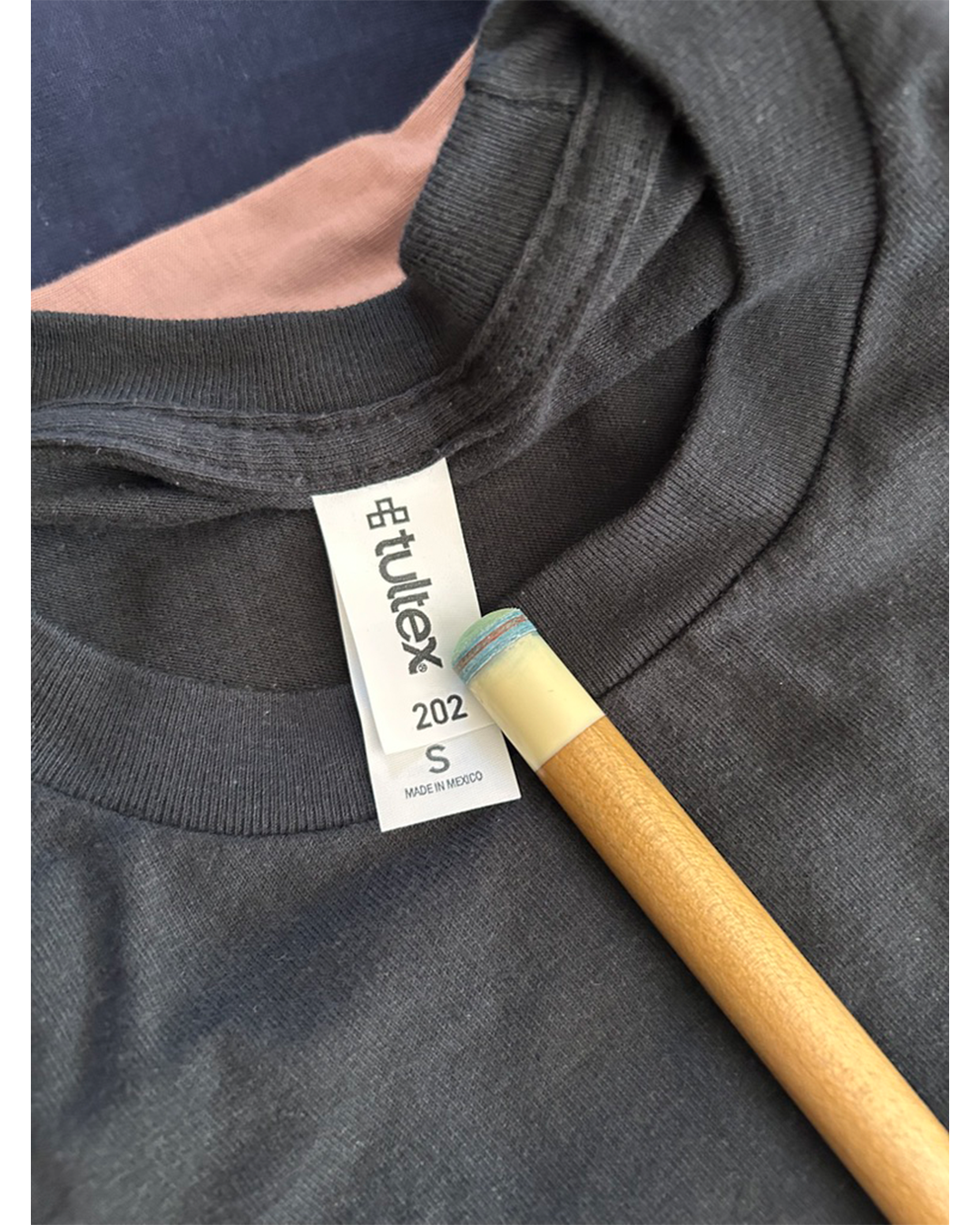 Close-up of a dark charcoal gray hoodie showing the inner neck label with the brand 'etulek' and size 'S'. A wooden stick with a blue and yellow eraser is resting against the collar.