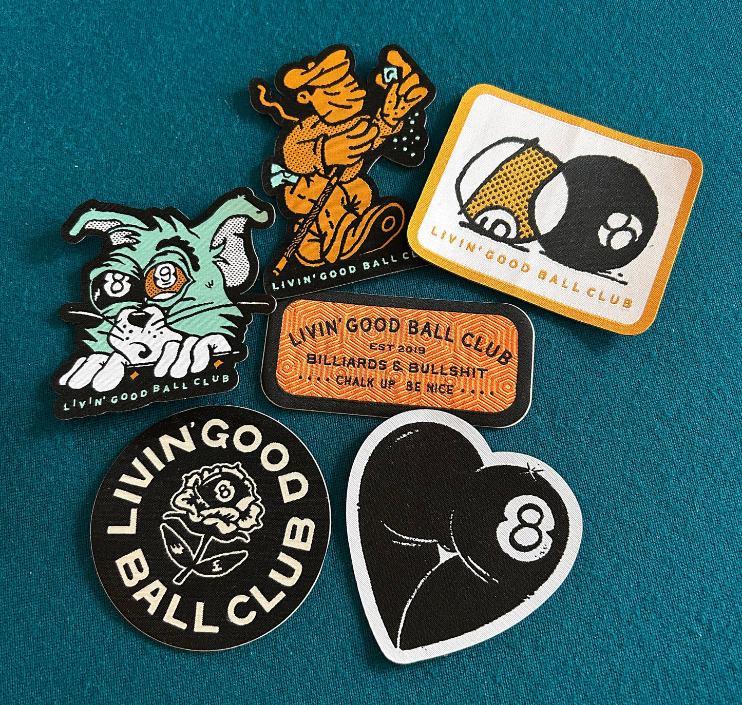 Five embroidered patches from the brand Livin' Good Ball Club arranged on a textured teal felt background. The patches feature various designs including a green cat, an orange dancing figure, a billiards scene, a black-and-white 8-ball heart, and a black-and-white rose logo patch.