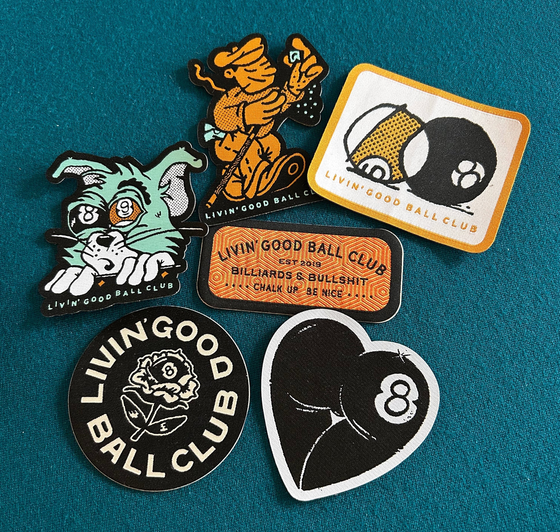 Five embroidered patches from the brand Livin' Good Ball Club arranged on a textured teal felt background. The patches feature various designs including a green cat, an orange dancing figure, a billiards scene, a black-and-white 8-ball heart, and a black-and-white rose logo patch.