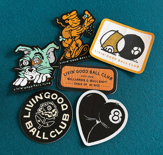 Five embroidered patches from the brand Livin' Good Ball Club arranged on a textured teal felt background. The patches feature various designs including a green cat, an orange dancing figure, a billiards scene, a black-and-white 8-ball heart, and a black-and-white rose logo patch.