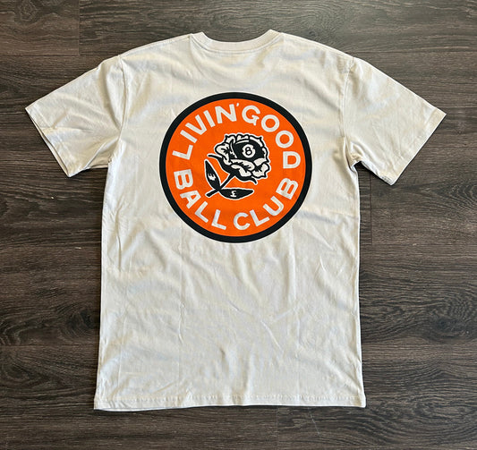 An off-white crewneck t-shirt laid flat on a dark wood floor, featuring a large circular graphic on the back with the text 'LIVING GOOD BALL CLUB' in white on an orange background with a black border.